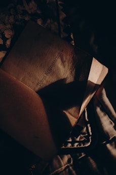 An open book with text on a page partially covered by shadows. The lighting is dim and creates a mysterious, intimate atmosphere, with fabrics underneath adding texture.