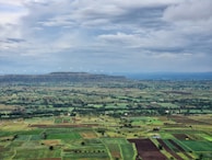 Expansive agricultural fields stretch across the landscape, showcasing various shades of green and brown. In the distance, a ridge is lined with wind turbines, set against a backdrop of a cloudy sky. The scene is peaceful and serene, with a mix of cultivated fields and patches of untouched land.