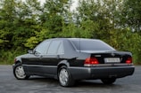 A classic black sedan is parked on an asphalt surface with lush green foliage in the background. The vehicle is well-kept, featuring a sleek design, tinted windows, and distinctive branding on the rear. The license plate is visible, adding to the detailed appearance of the car.