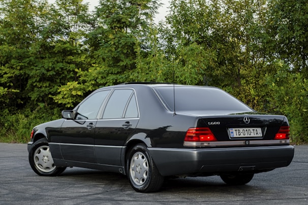 A classic black sedan is parked on an asphalt surface with lush green foliage in the background. The vehicle is well-kept, featuring a sleek design, tinted windows, and distinctive branding on the rear. The license plate is visible, adding to the detailed appearance of the car.