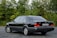 A classic black sedan is parked on an asphalt surface with lush green foliage in the background. The vehicle is well-kept, featuring a sleek design, tinted windows, and distinctive branding on the rear. The license plate is visible, adding to the detailed appearance of the car.