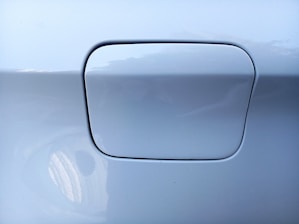 A close-up view of a car's fuel door with a smooth, reflective surface. The fuel door is part of a silver or light gray painted panel, and there are some subtle reflections visible.