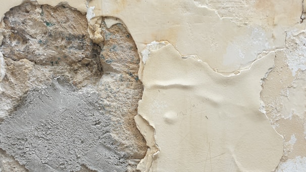 A close-up view of a wall with peeling paint and exposed plasterwork. The image shows various textures, including rough, uneven stonework and a smooth plaster surface that appears worn and cracked. The paint is chipped away in places, revealing multiple layers beneath.