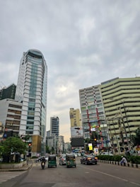 A busy urban scene with high-rise buildings surrounding a city intersection. The road is filled with vehicles, including cars and auto rickshaws, with some pedestrians visible on the sidewalk. The sky is overcast with gray clouds, and the buildings are adorned with various advertisements and signs.