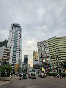 A busy urban scene with high-rise buildings surrounding a city intersection. The road is filled with vehicles, including cars and auto rickshaws, with some pedestrians visible on the sidewalk. The sky is overcast with gray clouds, and the buildings are adorned with various advertisements and signs.