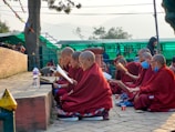 A group of people in red robes are seated outdoors, engaging in reading or study. They are positioned along a stone path, with trees and a green structure in the background. Some have shaved heads, and a couple are wearing blue face masks. They appear focused and intent on their task.