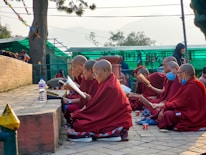 A group of people in red robes are seated outdoors, engaging in reading or study. They are positioned along a stone path, with trees and a green structure in the background. Some have shaved heads, and a couple are wearing blue face masks. They appear focused and intent on their task.