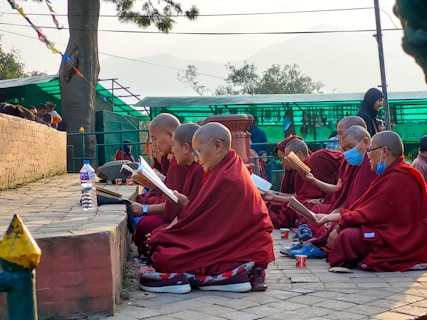 A group of people in red robes are seated outdoors, engaging in reading or study. They are positioned along a stone path, with trees and a green structure in the background. Some have shaved heads, and a couple are wearing blue face masks. They appear focused and intent on their task.