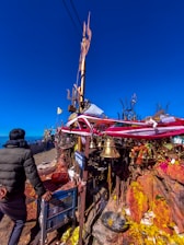 A person stands in front of a religious shrine adorned with colorful ribbons, golden bells, and flags. The shrine is set against a bright blue sky, with offerings and incense placed on a stone surface. Signs with text are visible, and there are various religious symbols and objects around the area.