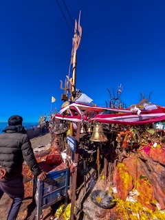 A person stands in front of a religious shrine adorned with colorful ribbons, golden bells, and flags. The shrine is set against a bright blue sky, with offerings and incense placed on a stone surface. Signs with text are visible, and there are various religious symbols and objects around the area.