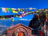 Brightly colored prayer flags flutter in the wind against a backdrop of snow-capped mountains and clear blue sky. A traditional brick shrine with intricate carvings stands in the foreground.