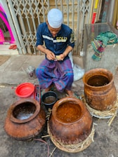 A person wearing a blue shirt and a white cap sits on the ground near several large clay pots filled with liquid. They appear to be counting paper currency while surrounded by various objects, including a red bowl, a metal bucket, and a bundle of colorful cloth on a metal cabinet next to them. The setting seems to be outdoors, possibly a street or market area.