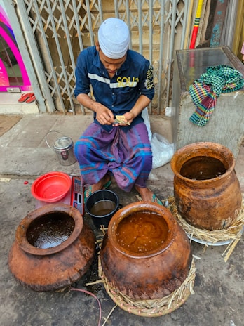 A person wearing a blue shirt and a white cap sits on the ground near several large clay pots filled with liquid. They appear to be counting paper currency while surrounded by various objects, including a red bowl, a metal bucket, and a bundle of colorful cloth on a metal cabinet next to them. The setting seems to be outdoors, possibly a street or market area.
