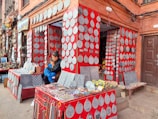 A street-side shop displays an array of intricately carved stone plaques, predominantly circular and teardrop in shape, mounted on vibrant red panels. The shop also offers various items for sale including bracelets and necklaces on a tabletop. A woman and child are seated together, the woman is engrossed in her mobile phone. The facade of the shop and surrounding area feature brickwork and a variety of artifacts and paintings.