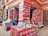 A street-side shop displays an array of intricately carved stone plaques, predominantly circular and teardrop in shape, mounted on vibrant red panels. The shop also offers various items for sale including bracelets and necklaces on a tabletop. A woman and child are seated together, the woman is engrossed in her mobile phone. The facade of the shop and surrounding area feature brickwork and a variety of artifacts and paintings.