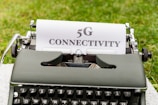 A vintage typewriter placed on grass has a sheet of paper with the text '5G CONNECTIVITY' typed in bold letters.