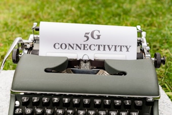 A vintage typewriter placed on grass has a sheet of paper with the text '5G CONNECTIVITY' typed in bold letters.