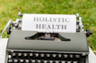 An old-fashioned typewriter displays a sheet of paper with the words 'Holistic Health' on it, set against a background of green grass. The typewriter features keys that are visible at the bottom of the image.