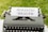 An old-fashioned typewriter displays a sheet of paper with the words 'Holistic Health' on it, set against a background of green grass. The typewriter features keys that are visible at the bottom of the image.
