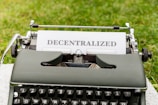 A vintage typewriter with the word 'DECENTRALIZED' typed on a sheet of paper. The typewriter is positioned outdoors on a grassy surface.