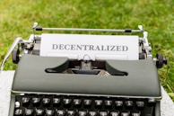 A vintage typewriter with the word 'DECENTRALIZED' typed on a sheet of paper. The typewriter is positioned outdoors on a grassy surface.