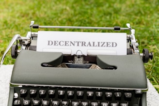 A vintage typewriter with the word 'DECENTRALIZED' typed on a sheet of paper. The typewriter is positioned outdoors on a grassy surface.