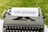 A vintage typewriter positioned outdoors on a grassy surface, with a sheet of paper in it displaying the word 'UPCYCLING' in bold letters.