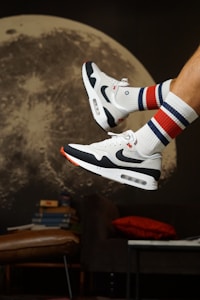 A pair of white and black sneakers with a swoosh logo is in mid-air against a backdrop of a large, detailed image of the moon. The sneakers are paired with white socks featuring blue and red stripes. In the background, there is a stack of books on a leather chair, a red cushion on a dark sofa, and part of a keyboard.