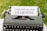 A vintage typewriter with a white paper inserted into it is displayed. The paper has the phrase 'INQUIRY-BASED LEARNING' typed in bold letters. The typewriter is set on a green grassy background.