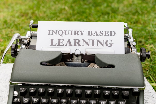 A vintage typewriter with a white paper inserted into it is displayed. The paper has the phrase 'INQUIRY-BASED LEARNING' typed in bold letters. The typewriter is set on a green grassy background.