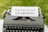 A vintage typewriter with a white paper inserted into it is displayed. The paper has the phrase 'INQUIRY-BASED LEARNING' typed in bold letters. The typewriter is set on a green grassy background.