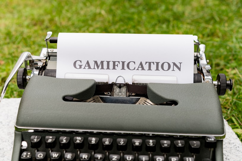 A vintage typewriter with a sheet of paper displaying the word 'GAMIFICATION' in bold letters. The scene is set against a backdrop of green grass, suggesting an outdoor environment.