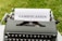 A vintage typewriter with a sheet of paper displaying the word 'GAMIFICATION' in bold letters. The scene is set against a backdrop of green grass, suggesting an outdoor environment.