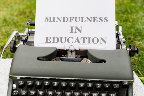 An old-fashioned typewriter set on a grassy surface holds a sheet of paper with the words 'Mindfulness In Education' typed on it. The typewriter features a dark green or gray color with classic keys displaying white letters.