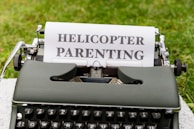 A vintage typewriter sits on a flat surface outdoors with a piece of paper inserted. The paper displays the bold text HELICOPTER PARENTING.