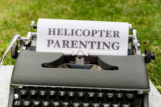A vintage typewriter sits on a flat surface outdoors with a piece of paper inserted. The paper displays the bold text HELICOPTER PARENTING.