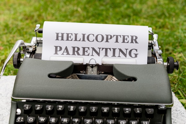 A vintage typewriter sits on a flat surface outdoors with a piece of paper inserted. The paper displays the bold text HELICOPTER PARENTING.