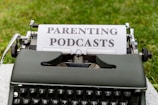 A vintage typewriter with a piece of paper in it, displaying the words 'Parenting Podcasts'. The typewriter is set on a grassy surface.