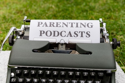 A vintage typewriter with a piece of paper in it, displaying the words 'Parenting Podcasts'. The typewriter is set on a grassy surface.