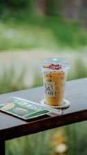 A cold beverage with ice and dark pearls is placed on a wooden table, next to a wrapped snack with green packaging. The background is a blurred view of greenery.