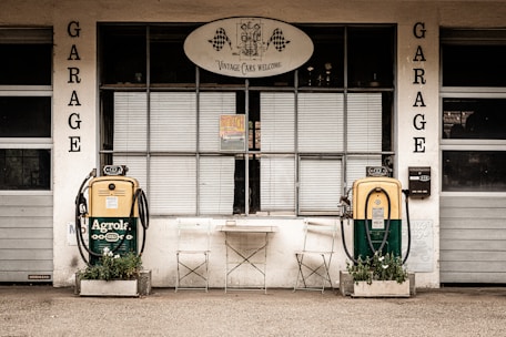A vintage garage facade featuring two retro gas pumps labeled 'Agrolal', with a sign above the garage reading 'Vintage Cars Welcome'. The gas pumps are yellow and green, surrounded by small planters with greenery. Two folding metal chairs and a small table are positioned between the pumps, suggesting a waiting area. The garage door and windows have a rustic, aged appearance.