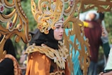 A person dressed in an ornate costume featuring detailed gold and red patterns, with a large headpiece consisting of intricate designs and embellishments. The costume includes bold makeup and striking facial adornments, creating an elegant and dramatic look. In the background, other people are slightly blurred, focusing attention on the central figure.