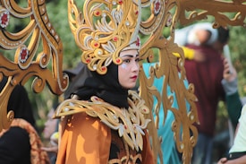 A person dressed in an ornate costume featuring detailed gold and red patterns, with a large headpiece consisting of intricate designs and embellishments. The costume includes bold makeup and striking facial adornments, creating an elegant and dramatic look. In the background, other people are slightly blurred, focusing attention on the central figure.