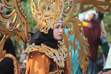 A person dressed in an ornate costume featuring detailed gold and red patterns, with a large headpiece consisting of intricate designs and embellishments. The costume includes bold makeup and striking facial adornments, creating an elegant and dramatic look. In the background, other people are slightly blurred, focusing attention on the central figure.