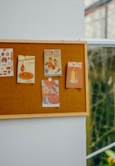 A bulletin board mounted on a wall displays several vintage-style advertisements and posters related to food items. The posters include images of pancakes, chocolate, and other food products pinned on the board. The board is positioned next to a window, allowing natural light to illuminate the posters.