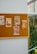 A bulletin board mounted on a wall displays several vintage-style advertisements and posters related to food items. The posters include images of pancakes, chocolate, and other food products pinned on the board. The board is positioned next to a window, allowing natural light to illuminate the posters.