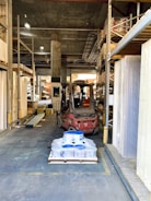 A warehouse aisle containing metal shelves filled with various building materials. In the center is a forklift, which appears to be moving a pallet loaded with blue and white containers. The surroundings are industrial, with visible shelving, concrete walls, and metal beams. The space has a cluttered yet organized feel, typical of large storage facilities.