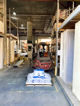 A warehouse aisle containing metal shelves filled with various building materials. In the center is a forklift, which appears to be moving a pallet loaded with blue and white containers. The surroundings are industrial, with visible shelving, concrete walls, and metal beams. The space has a cluttered yet organized feel, typical of large storage facilities.