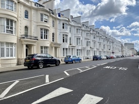 A row of elegant, white, terraced houses lines a street with several parked cars, including a black SUV and a blue car. The architecture features large windows and ornate details, with a clear blue sky and scattered clouds above.