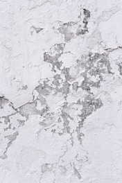 Cracked and peeling white paint on a textured surface reveals patches of dull gray material beneath. The uneven layers create a rugged and weathered appearance.